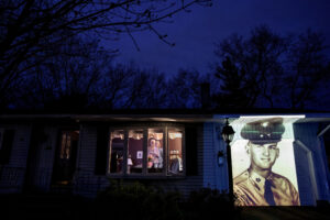 An image of veteran Francis Foley is projected onto the home of his wife, Dale Foley, left, as she looks out a window with their daughter, Keri Rutherford, in Chicopee, Mass., Wednesday, April 29, 2020. Foley, a U.S. Army veteran and resident of the Soldier's Home in Holyoke, Mass., died from the COVID-19 virus at the age of 84. Foley never learned to read music but could play any song by ear. He loved a cup of coffee and something sweet from Dunkin’ Donuts. He kept the nurses at the home laughing. He was fiercely protective of his family. Ask his family about the man they lost, and the words flow easily about the card-carrying union carpenter, Army veteran, devoted husband of 54 years and father of four. “He was strong. He was funny. He was engaging. He was ornery. He was feisty,” his daughter, Keri, says. “He was still full of life. And then within days, he’s gone.”