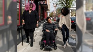 Three Black and disabled folx smile and stroll down a sidewalk side by side. On the left, a non-binary person walks with a cane in one hand and a tangle stim toy in the other. In the middle, a non-binary person rolls along in their power wheelchair. On the right, a femme gestures mid-walk. Photo Credit: Disabled And Here