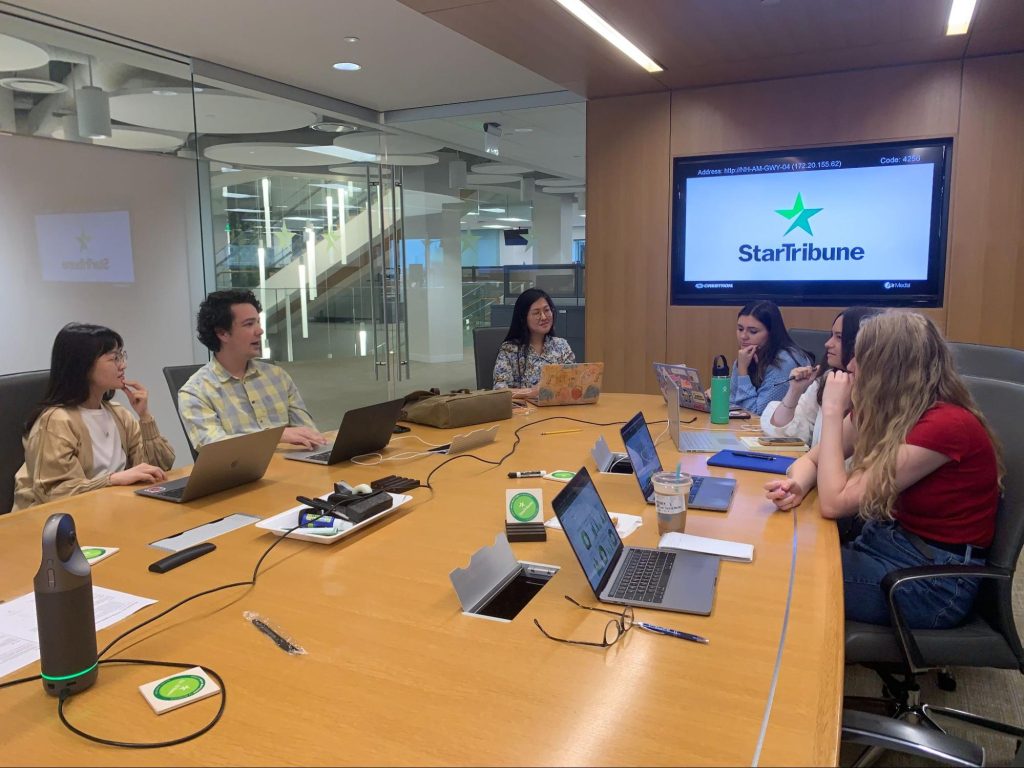 Six interns sit around a large oval table with their laptops out, having a conversation. A TV in the background showcases the Star Tribune logo. The newsroom is visible through the glass doors in the corner.