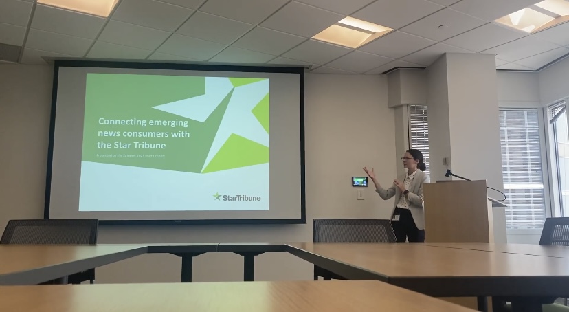 Sydney stands to the right of a large projector screen in a conference room, gesturing to a slide that reads, "Connecting emerging news consumers with the Star Tribune"