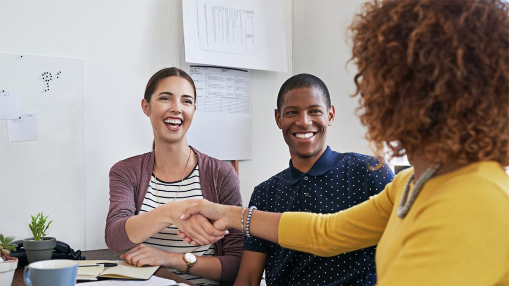 Two women shake hands while a smiling man looks on