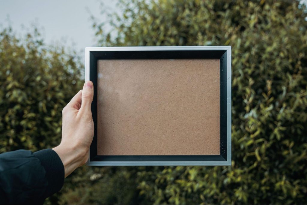 A hand holds up an empty picture frame in front of a blurry background of bushes and trees.