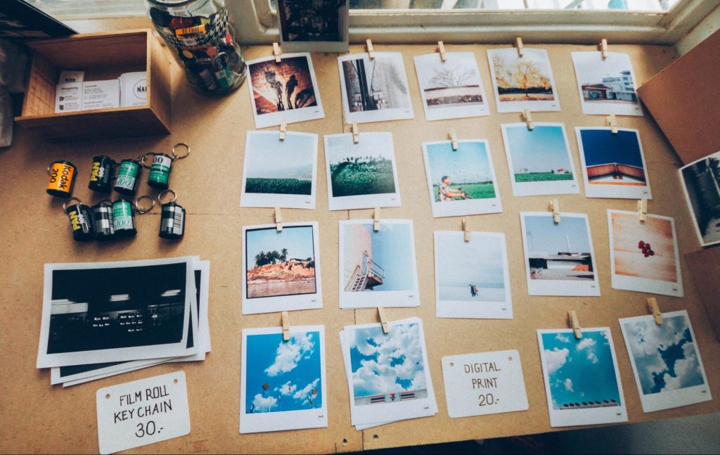 A large desk has many printed photos laid out on it, each depicting a different scene. Several rolls of film and a stack of unsorted photos rest on the desk which is illuminated by light from a window.