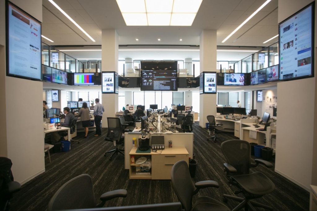 A newspaper newsroom with many desks in an open "bullpen" style room, empty rolling desk chairs and some people standing and sitting at desks, digital monitors hang from the walls