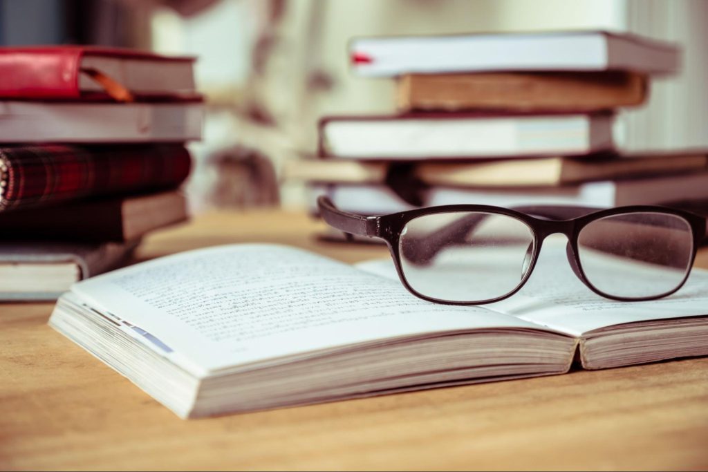 Close up of open book with eyeglasses on wooden desk, with a variety of different books stacked on top of each other in the background.