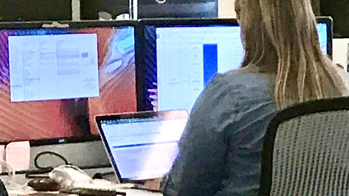 Journalist sits at desk with laptop and multiple monitors.