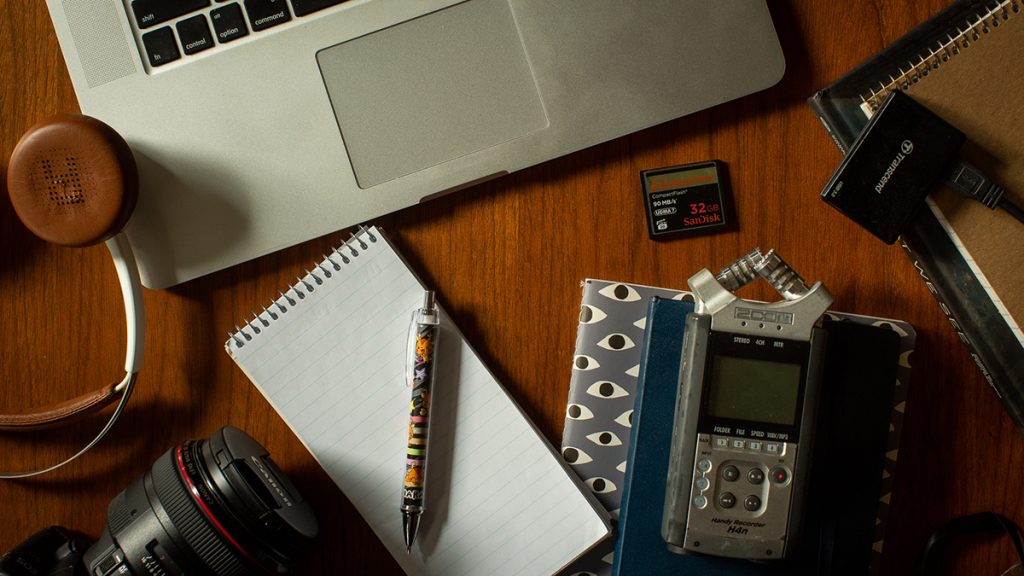 A desktop with reporter tools, including. laptop, headphones, a camera, a notepad, a journal, an audio recorder and a microphone. Photo: Michelle Kanaar