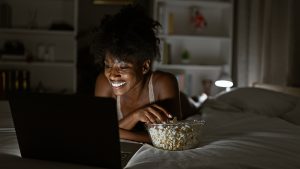 Smiling woman lying on a bed watching something on a laptop while eating popcorn