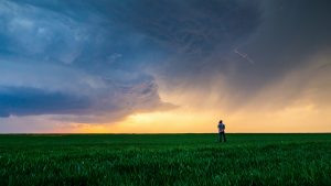 Person stands in a field with a camera on a tripod photographing a massive storm cloud formation.