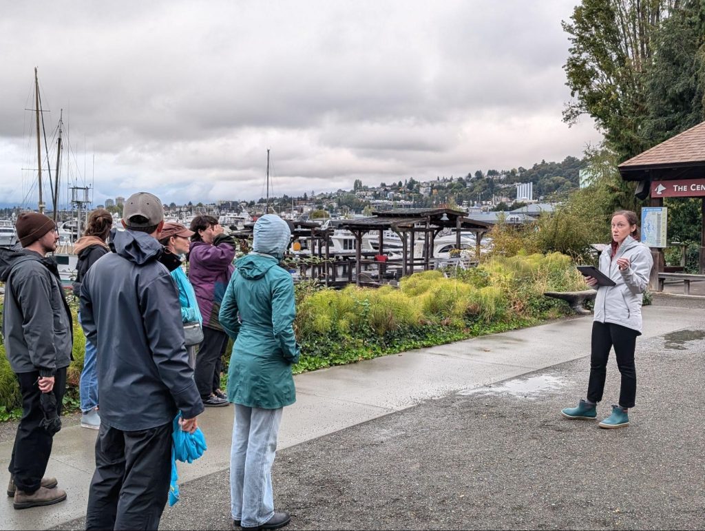 Cara Kuhlman guides a walking tour in Seattle. The group stands next to boat docks on an overcast day