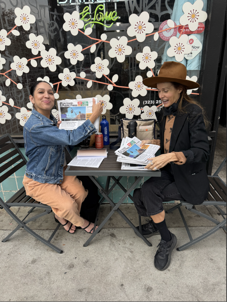 Two women at a café table with multiple copies of LA Public Press's newspaper