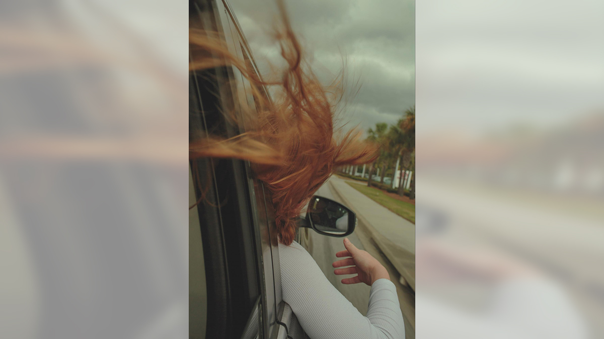 Redheaded woman's long hair blowing behind her as she puts her arm and head out of the passenger side window of a car going down a rural road.