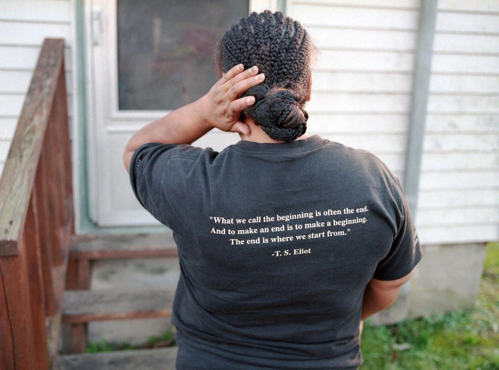 A woman with braided locks gathered into a bun touches her hair. The back of her t-shirt has a quote from T.S. Eliot, "What we call the beginning is often the end. And to make an end is to make a beginning. The end is where we start from."