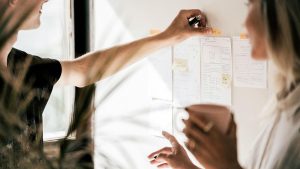Woman tapes a label to a whiteboard with several pieces of paper taped to it, while another woman looks on.