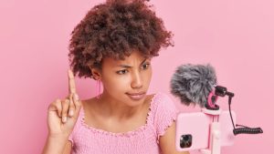 Serious attentive Afro American woman raises index finger focused at smartphone camera has online conversation with subscribers gives important tips isolated over pink background. Video blogging