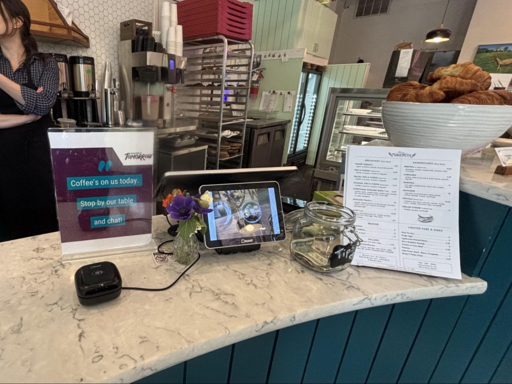 A coffee shop counter with a sign, "Coffee's on use today. Stop by our table and chat."