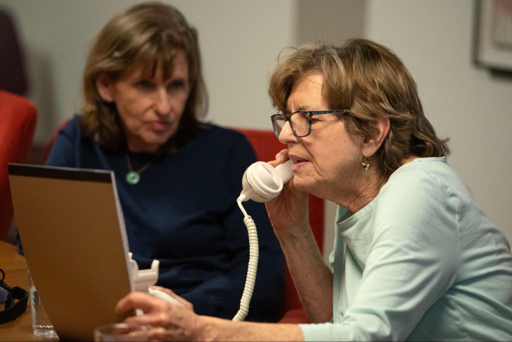 Two women look at a notebook while one talks on the telephone.
