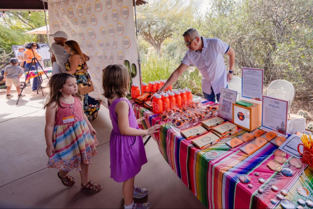 Two girls look over items a man has for sale on a table at an event
